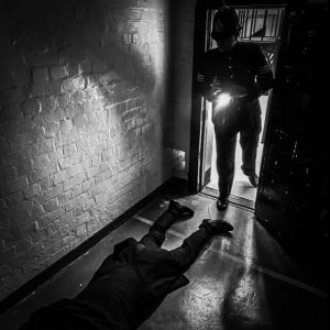 black and white image of a policeman standing in a door way, his torch shining on a dead body