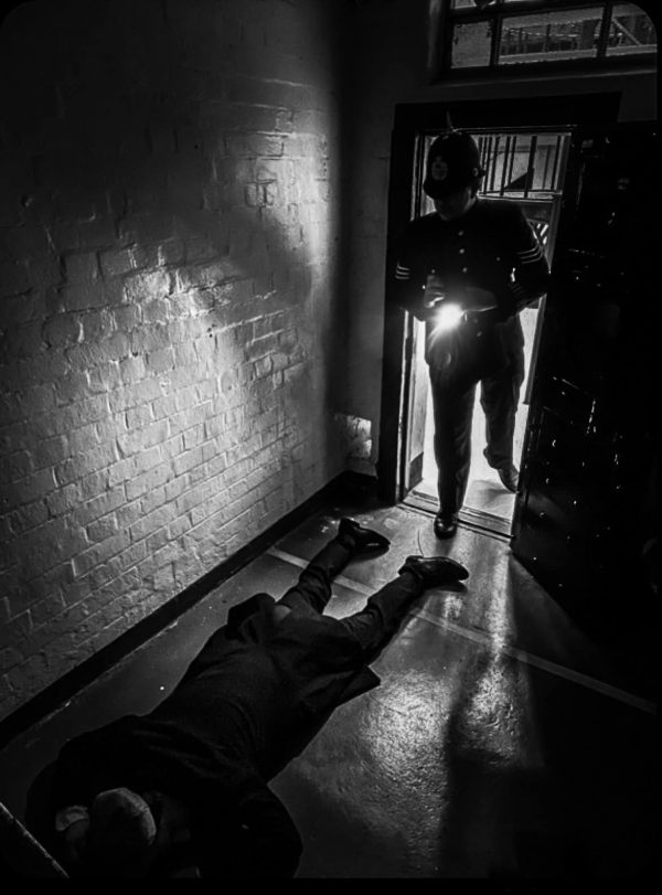 black and white image of a policeman standing in a door way, his torch shining on a dead body