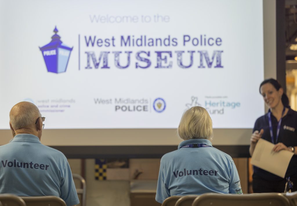two volunteers in blue polo shirts sit in front of a projector image reading West Midlands Police Museum