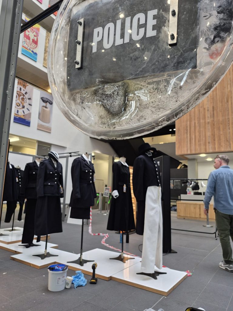 a damaged police riot shield is on display. In the background several police uniforms are being put on mannequins