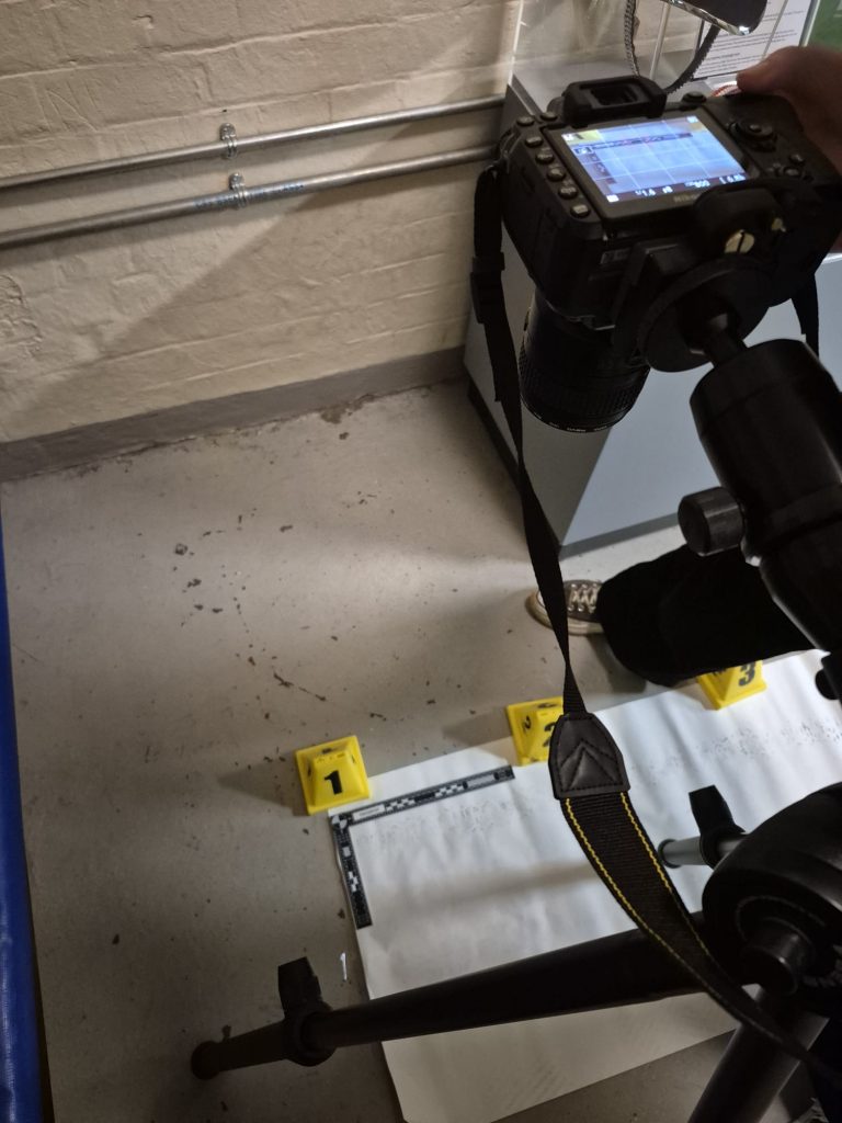 a digital camera faces the floor, capturing a photo of show prints. Next to the shoe prints is a ruler and yellow numbered cones.
