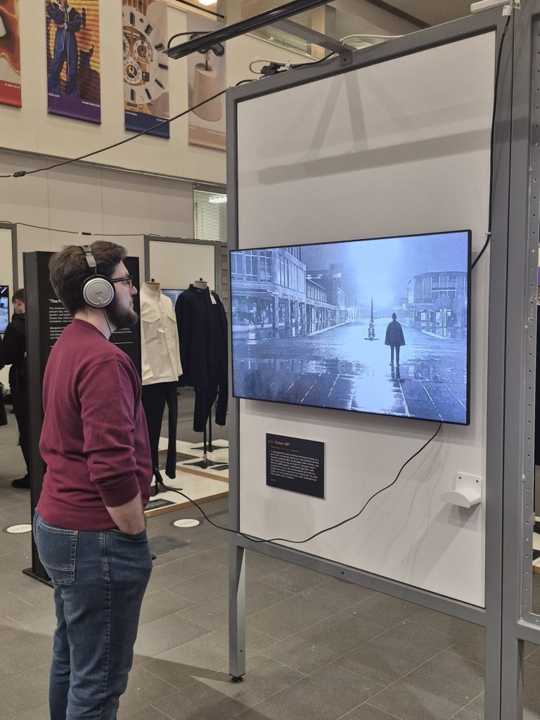 A young man is wearing a set of headphones connected to a tv. The tv shows an image of a police officer in the 1960s.